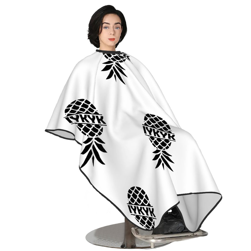 White hair cutting cape with upside down pineapple with IYKYK on it. Swinger merchandise. Woman Sitting in chair with white hair cutting cape with upside down pineapple on it.