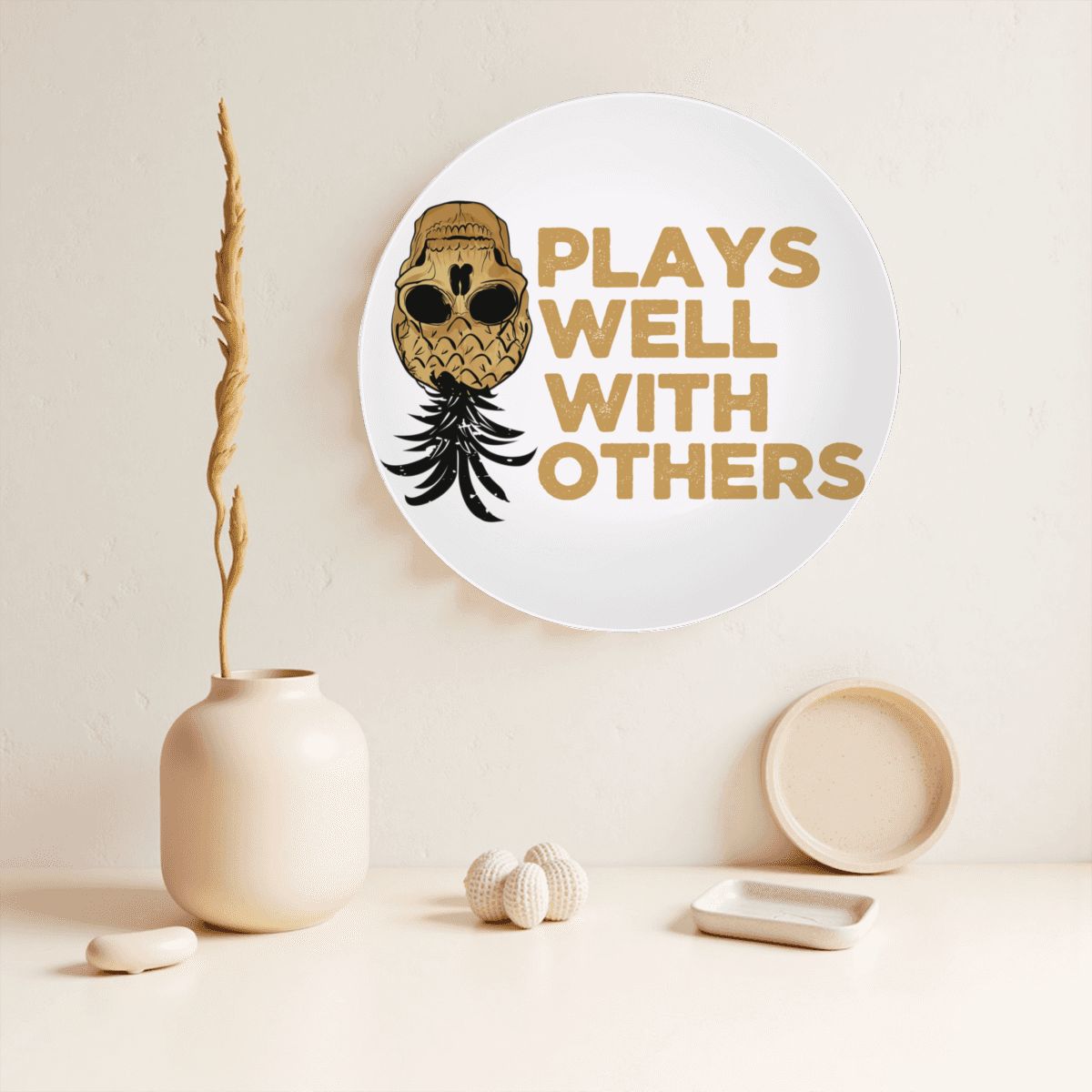 Plays Well With Others decorative plate hanging on a beige wall next to a vase and other house hold good idems.