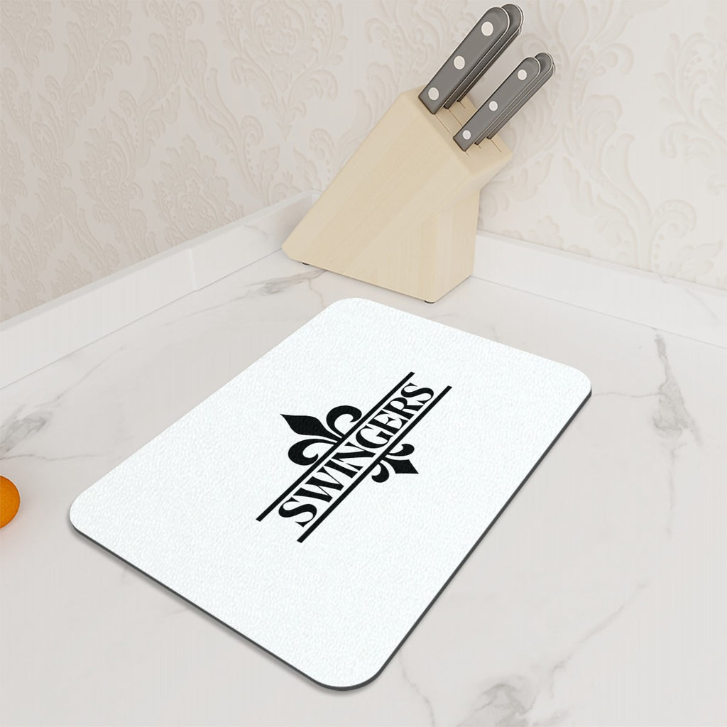 Glass cutting board with a Fluer-di-lis with Swingers in the middle of it sitting on a counter op next to a group of knives.