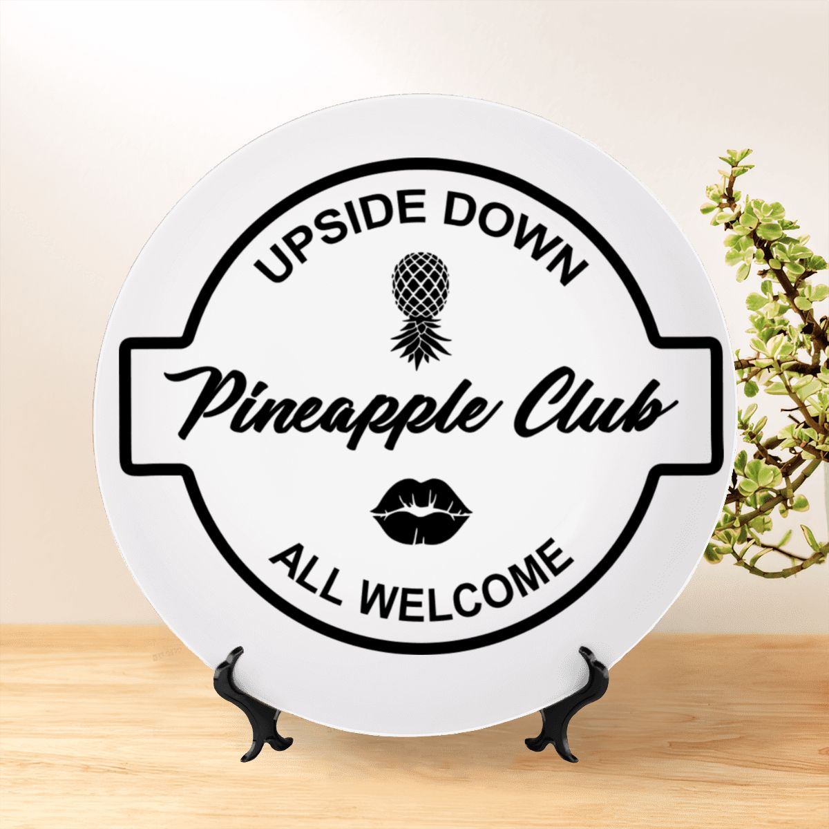 Upside-Down Pineapple Club decorative plate on a wooden shelf next to a plant in a home. 